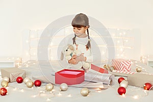 Charming little girl wearing white sweater embracing soft toy dog, sitting on bed with Christmas decoration and garland, looking