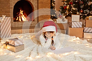 Charming little girl wearing white sweater and santa claus hat, lying on floor near Christmas tree, present boxes and fireplace,