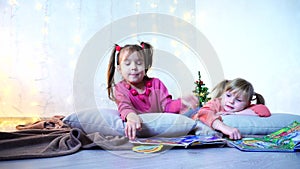 Charming little girls play together and chat, lying on floor and on pillows against wall with garland and Christmas tree