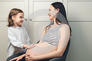 Charming little teenage girl holds her hand on her mother's pregnant belly. Pregnant woman with her little daughter at home.