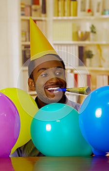 Charming man wearing blue shirt and hat sitting by table with balloons blowing party horn celebrating alone smiling