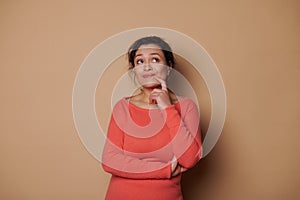 Charming middle-aged woman, thoughtfully looking aside, holding finger near her lips while reasoning on beige background