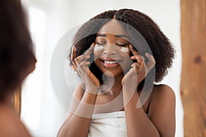 Charming millennial Afro lady applying nourishing facial cream under her eyes near mirror at home