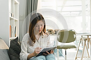 Charming millennial Asian woman is using her digital tablet while relaxing on the sofa at home.
