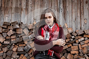 Charming model of a pretty young woman in a stylish knitted sweater with a fashionable scarf stands near an old wooden wall with