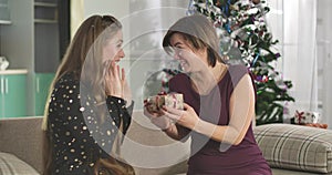 Charming mother giving Christmas gift to adult daughter. Portrait of happy family celebrating New Year eve at home