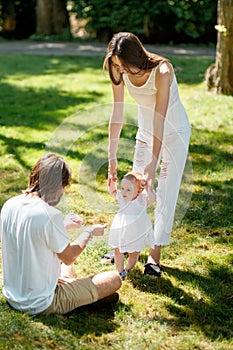 Charming mother and happy dad are teaching their little daughter wearing white dress how to make her first steps.