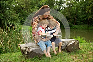 Charming Mother Reading a Book to Her Adorable Little Twin Sons While Sitting Outside Near Beautiful Lake