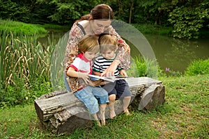 Charming Mother Reading a Book to Her Adorable Little Twin Sons While Sitting Outside Near Beautiful Lake