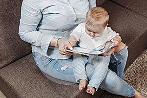 Charming mother showing images in a book to her cute little son at home sitting on the couch