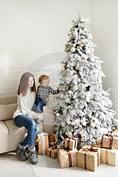 Charming mother in the white sweaterholding her little toddler son near Christmas tree and gift boxes in living room in the house