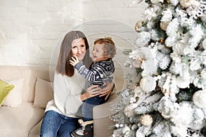Charming mother in the white sweaterholding her little toddler son near Christmas tree in the house