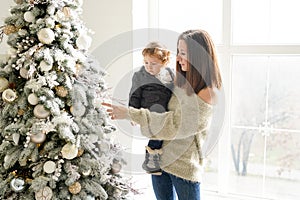 Charming mother in the white sweaterholding her little toddler son near Christmas tree in the house