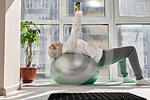 Charming Muslim woman cute smiling at the camera, doing exercises on the hands with dumbbells while lying on a sports ball