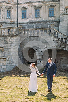 Charming newlywed bride and groom holding hands on green lawn near beautiful ruined baroque palace