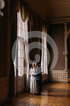Charming newlywed pair posing near large illuminated window in vintage victorian mansion