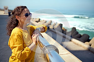 Charming pensive Hispanic woman in sunglasses, looking into the distance, standing against Atlantic Ocean background