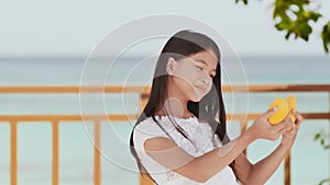 A charming philippine schoolgirl girl in a white dress and long hair positively poses with a mango in her hands. The sun