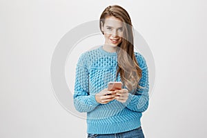 Charming positive fashion girl holding smartphone while looking ans smiling broadly at camera, typing something over