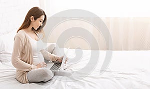 Charming pregnant woman relaxing with her laptop on bed