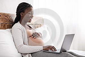 Charming pregnant woman relaxing with her laptop, browsing or chatting with friends online, sitting on bed, panorama