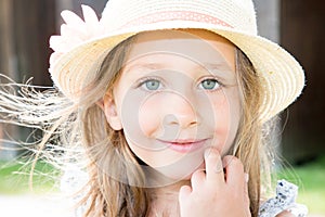 Charming preschool girl with big blue eyes and long loose blonde hair wearing straw hat looking at camera with hand finger on her