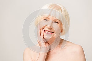 Charming, pretty, old woman touching her perfect soft face skin with fingers, smiling at camera over gray background, using day,