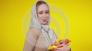 Charming redhead slim woman with green eyes in kerchief posing with poultry eggs at yellow background. Smiling beautiful
