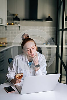 Charming redhead young woman having video call conversation using laptop and drinking champagne.