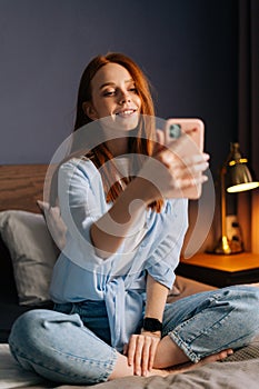 Charming redhead young woman taking mobile selfie photo on cellphone at cozy bedroom.