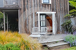 Charming scene - Two weathered wooden chairs on porch outside open door to bedroom in rustic house surrounded by plants
