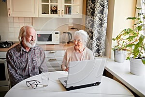 Charming senior couple using laptop sitting at the table in the kitchen room.