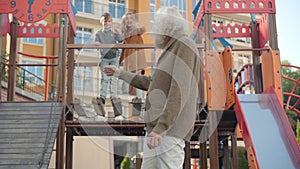 Charming senior man waving back to children having fun on playground and crossing hands. Portrait of happy old Caucasian