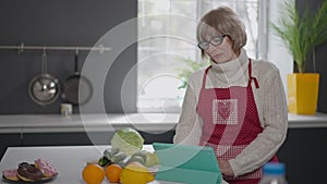 Charming senior woman surfing Internet on tablet standing in kitchen at home indoors. Portrait of confident beautiful