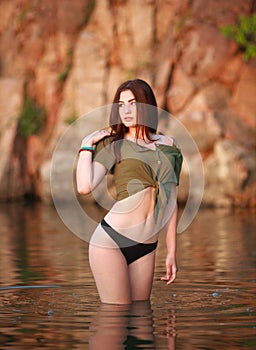 Charming sensual carefree brunette girl in water of wild lake on rock background in light of setting sun