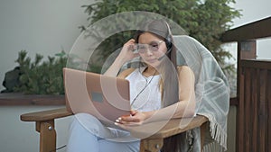 Charming serious Asian woman in eyeglasses and earphones sitting on armchair with laptop thinking. Portrait of confident