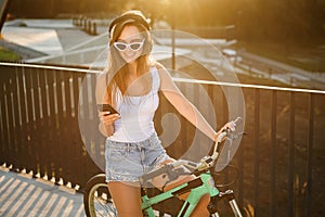 Charming sexy girl in sunglasses and tempting clothes sits on bicycle and listens to music with earphones on the sunset