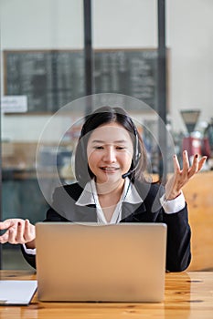 Charming and smiling Asian female customer service worker wearing headset in online meeting office look at laptop computer screen,