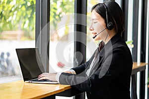 Charming and smiling Asian female customer service worker wearing headset in online meeting office look at laptop computer screen,