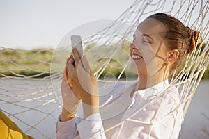 Charming smiling Caucasian woman wearing white shirt sitting in hammock on the bank of the river and using cell phone, typing