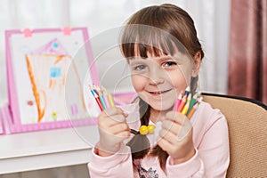 Charming smiling happy female child sitting with many colored pencils in both hands , looks at camera, finished her picture, kid