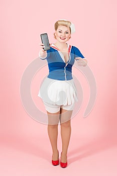 Charming stylish sailor female showing black touch screen of smart phone to the camera and pointing with finger on gadget.