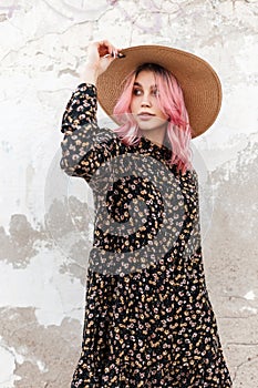 Charming stylish young woman with luxurious pink hair in trendy black floral dress in fashion straw hat posing near vintage wall