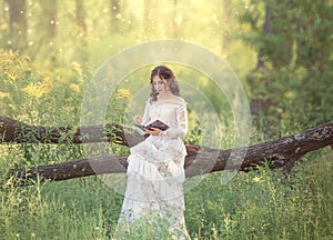 Charming sweet girl with dark hair and bare shoulders in a gorgeous vintage white dress sits on a fallen tree and reads