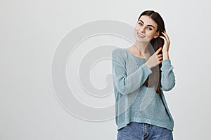 Charming and tender girl with sensual smile touching hair with both hands while standing over gray background and
