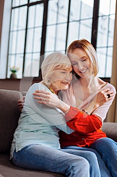Charming woman affectionately cuddling elderly madam sitting on couch