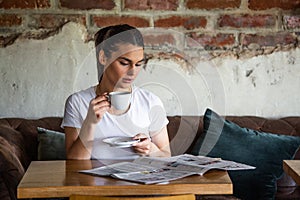 Charming woman with beautiful smile reading newspaper during rest in coffee shop, happy Caucasian female reading news while