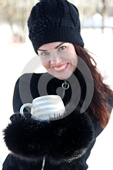 A charming woman in a black knitted hat with a pompom holds a beige striped Cup in black fur mittens, selective focus on