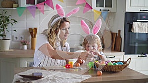 charming woman and her two-year-old son in bunny ears paint Easter eggs with paints sitting at the table on the kitchen