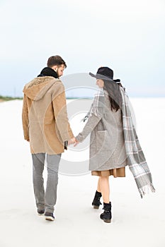 Charming woman and man walking on snow and holding hands, wearing coat, grey scarf and hat.